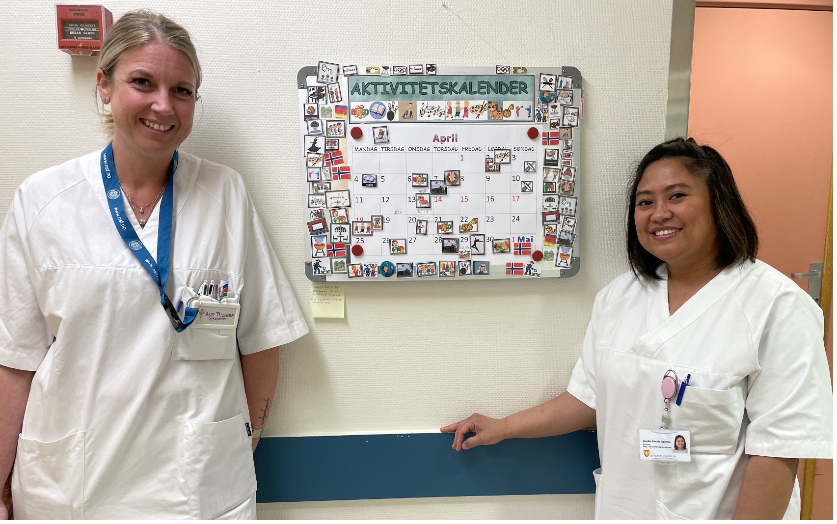 To kvinner i hvite sykepleier-frakker står foran en kalender som viser hva slags aktiviteter sykehjemmet skal ha denne måneden.